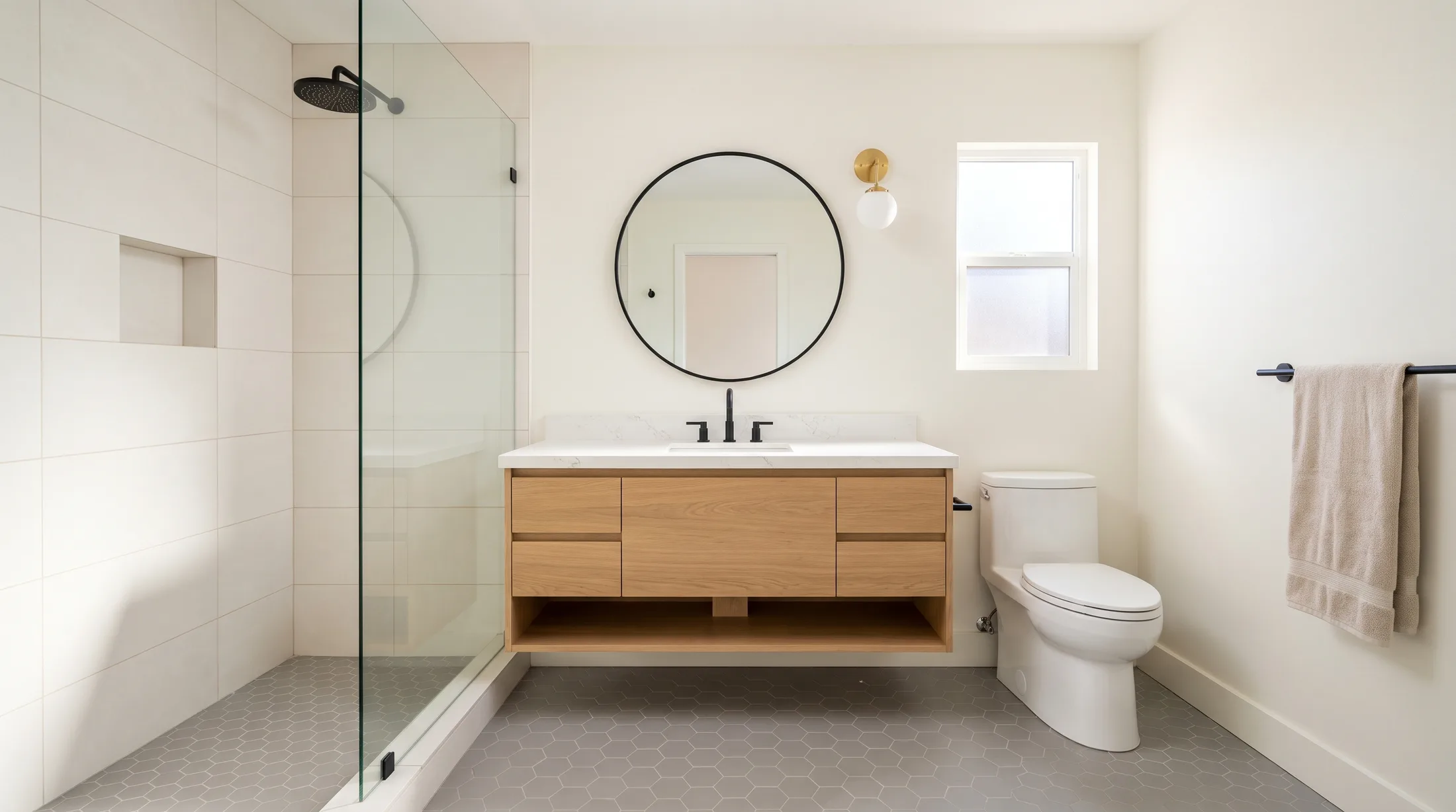 The bathroom finished: the frameless glass shower, the oak vanity under a round mirror, the brass sconce and a towel on the bar